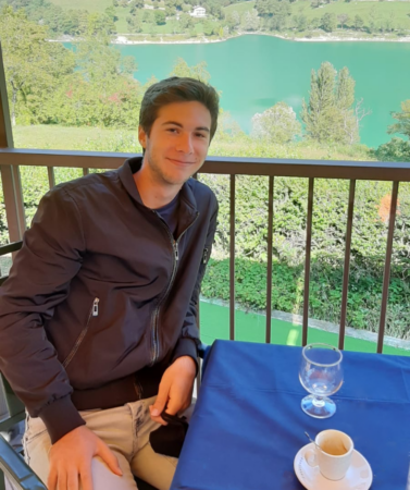 Matteo seduto a un tavolino con alle spalle un panorama naturale verdeggiante 